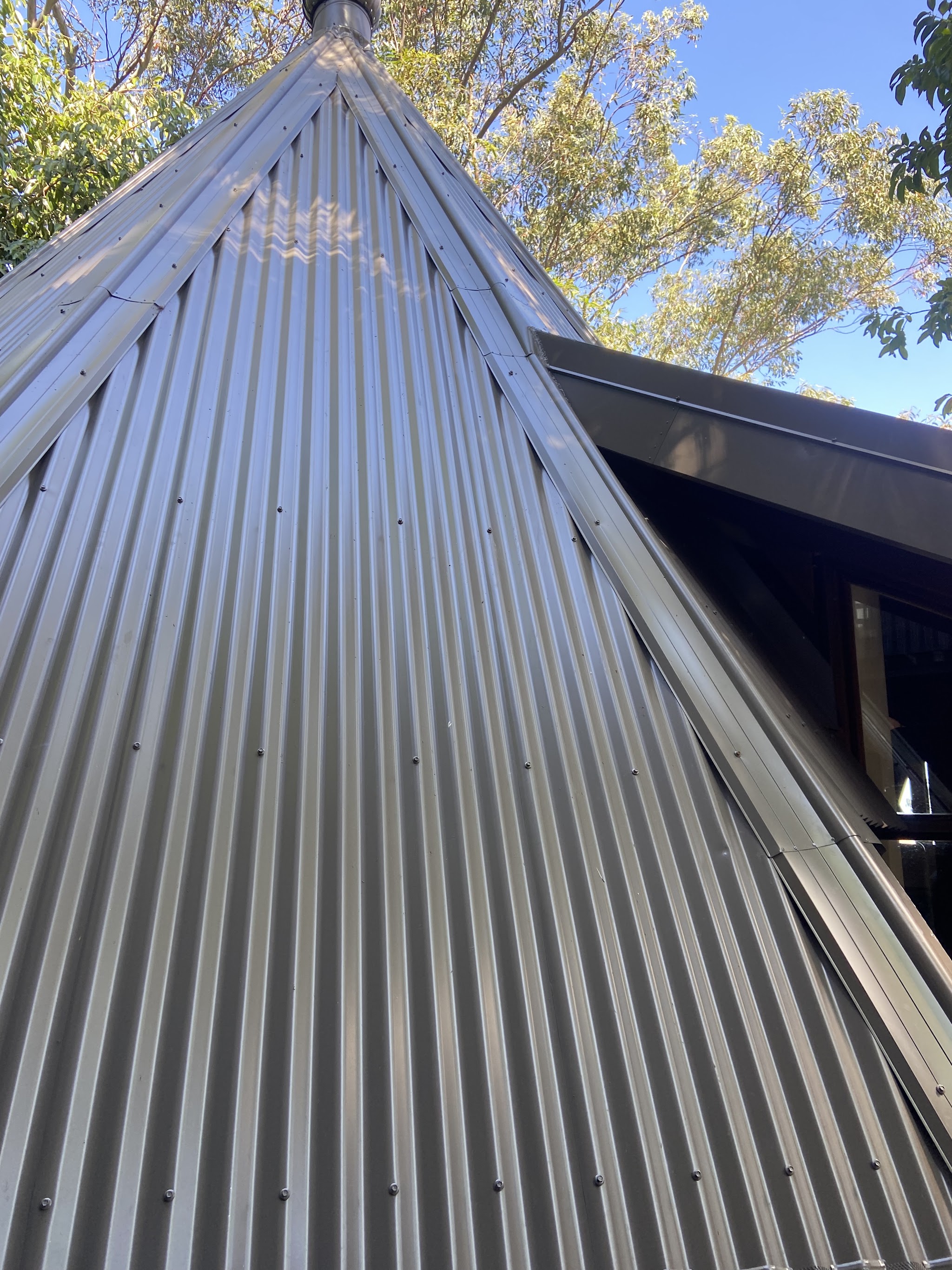 Looking up the ridge of a steep corrugated metal roof against blue sky and gum trees